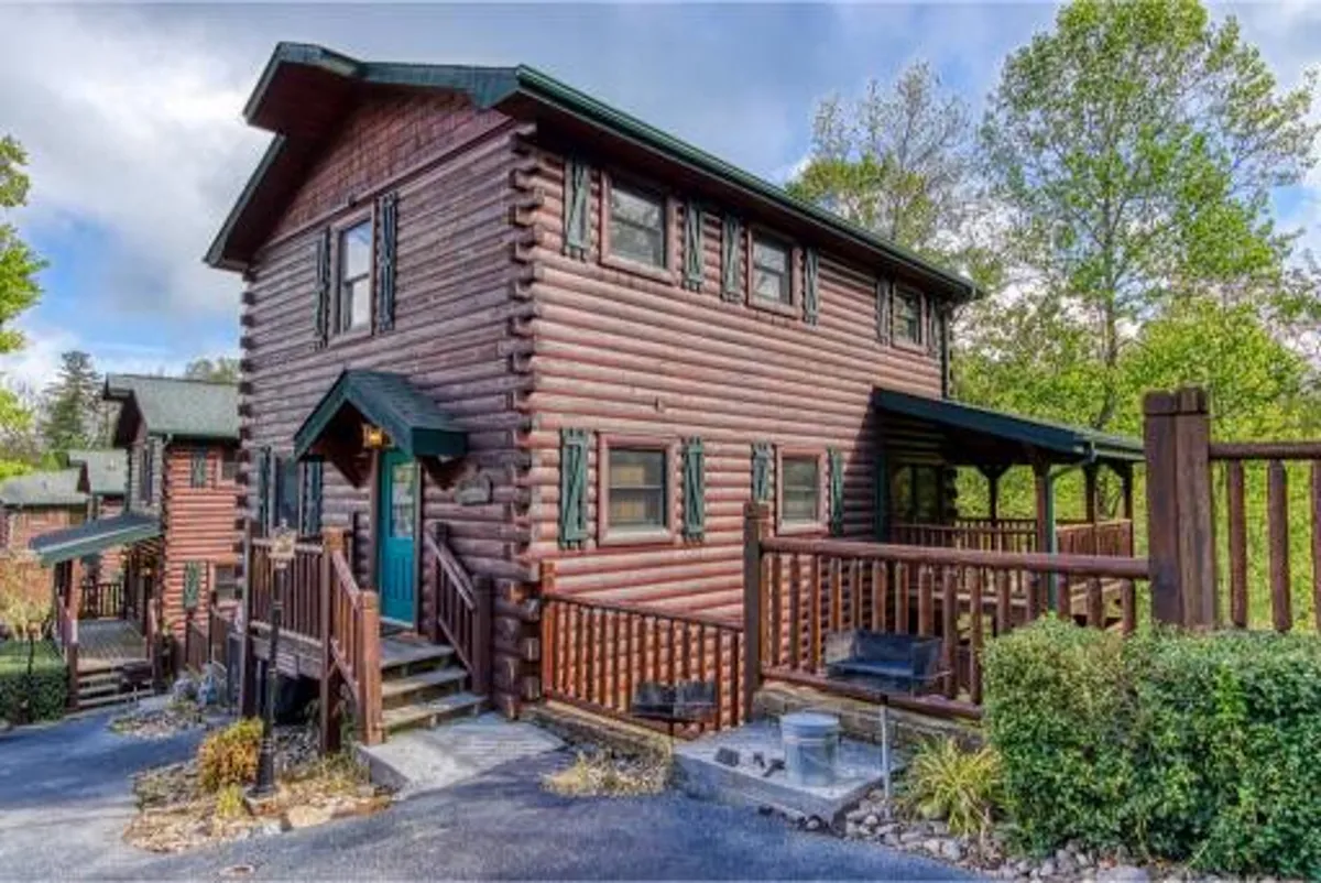 The image showcases the inviting exterior of a luxury log cabin with charming green shutters and an expansive wooden deck. The setting is surrounded by lush Tennessee forest, with the soft glow of sunset casting a warm, golden light across the deck—an ideal spot for gathering with loved ones or savoring a peaceful morning coffee immersed in nature.