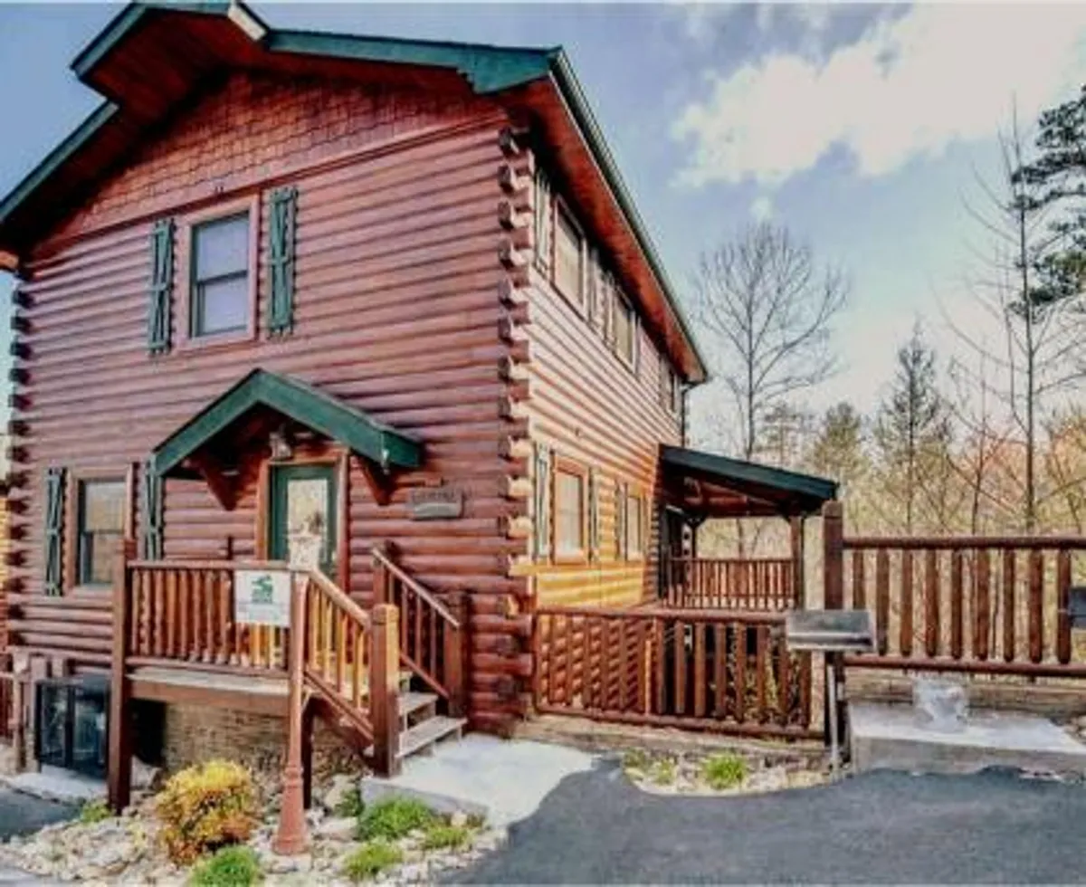 This image displays the welcoming exterior of a log cabin with green shutters and a wooden railing, surrounded by lush greenery. The sunset enhances the inviting mood, and a fire pit area suggests a spot for evening gatherings.