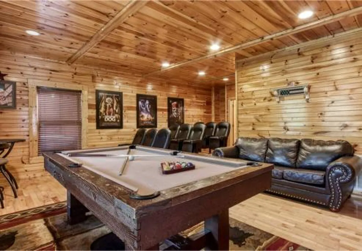 A cozy game room featuring a classic pool table at the center, a plush leather couch positioned nearby, wood-paneled walls, and artwork that adds warmth and character. The layout invites group play and relaxed socializing, with the elements of the room emphasizing both competition and comfort.