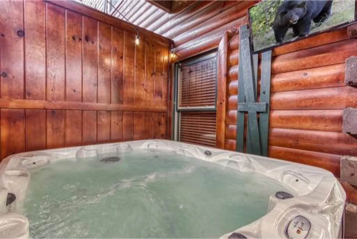 An indoor hot tub area with wooden walls, a decorative bear picture, and a window offering glimpses of the outdoors. The bubbling tub is the focal point, embodying indoor luxury while retaining a rustic cabin feel.
