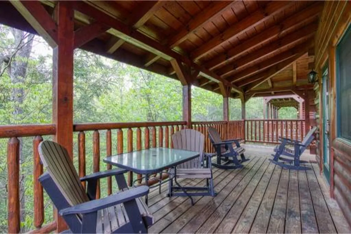 An inviting exterior porch scene featuring wooden rocking chairs and a glass table, bordered by a wooden railing and surrounded by lush greenery. The setup suggests a peaceful retreat where guests can unwind and connect with nature, perhaps enjoying coffee or quiet moments with friends.