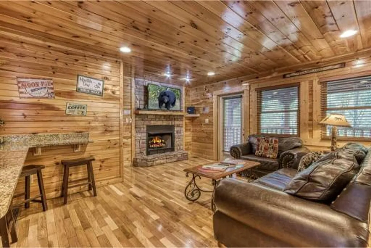 The image features a warm, open-concept living area with a central stone fireplace, a plush sofa, a television, and a bar area, all framed by natural wooden walls and plenty of natural light. The arrangement encourages relaxation and social gatherings, inviting guests to cozy up by the fire or enjoy entertainment together.