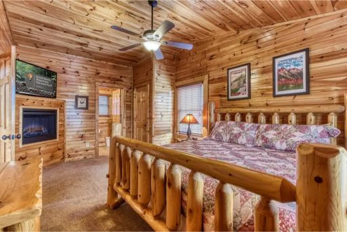A warmly lit bedroom featuring a substantial log bed with plush bedding, a rustic fireplace for ambiance, a ceiling fan, and a bedside lamp. The wooden walls and cabin details evoke a cozy, inviting atmosphere ideal for relaxation.