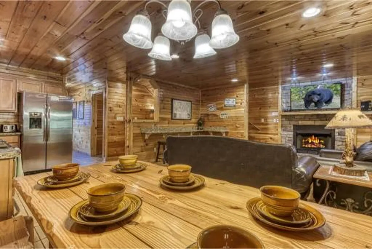 A warm, inviting dining area with a beautifully set table, chandelier lighting overhead, a cozy fireplace with a television above, and wooden cabinetry in the background. The space is suited to both intimate family dinners and festive celebrations.
